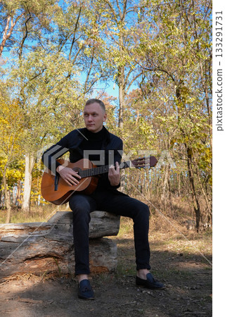 Beautiful blonde man playing acoustic guitar and singing relaxing enjoying outdoors on sunny autumn day. Artist learning and practicing playing musical instrument. Fall leaves autumnal 133291731