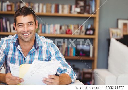Man in plaid shirt holding printed documents, smiling on couch in home office, copy space 133291751
