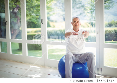 Senior man balancing on blue stability ball in home gym by white-framed glass doors, copy space 133291757