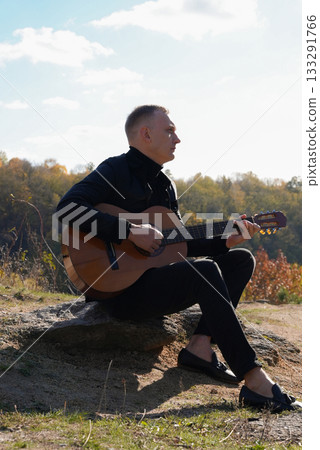 Portrait of smiling caucasian artist plays wooden acoustic guitar in autumnal park. Young songwriter plays string musical instrument hobby outside in nature fall time. Audio music healing 133291766
