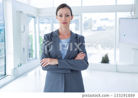 Female executive wearing grey blazer standing arms crossed in lobby near glass walls, screen, plant 133291889