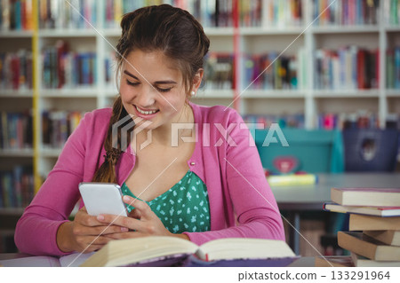 Teenage student smiling scrolling smartphone beside open book on library table bookshelves, bins 133291964