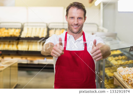 Mature adult man wearing red apron giving thumbs up behind bakery counter with bread and pastries 133291974