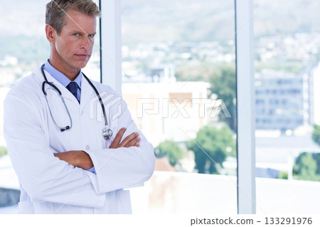 Male doctor standing with arms crossed, lab coat and stethoscope in office viewing city, copy space 133291976
