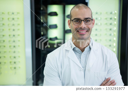 Smiling male optometrist donning lab coat in optical shop with lit display panels, eyeglass frames 133291977