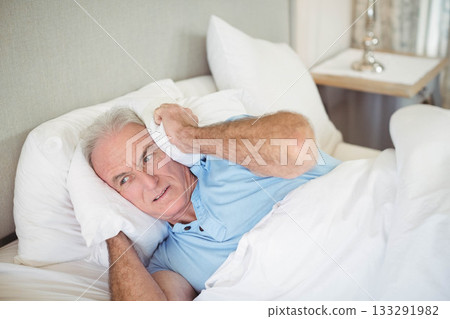 Senior man holding pillow against ear while lying in bedroom wearing light blue polo under duvet 133291982