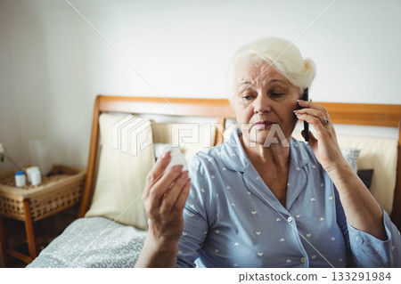 Senior woman reading pill bottle and holding smartphone while sitting on bed in bedroom, copy space 133291984