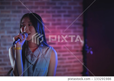 Asian woman singing into microphone on stand on club stage wearing sleeveless denim top, copy space Asian woman singing into microphone on stand on club stage wearing sleeveless denim top, copy space 133291986
