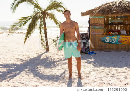 Shirtless man standing barefoot on beach holding surfboard beside surf rental hut with chalk sign 133291990