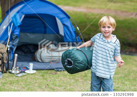 Boy carrying rolled sleeping bag while standing on grassy campsite by blue dome tent, copy space Boy carrying rolled sleeping bag while standing on grassy campsite by blue dome tent, copy space 133291998