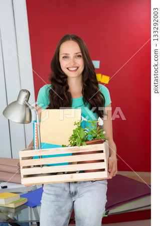 Woman smiling while holding crate filled with file folders, potted plant and desk lamp in office Woman smiling while holding crate filled with file folders, potted plant and desk lamp in office 133292003