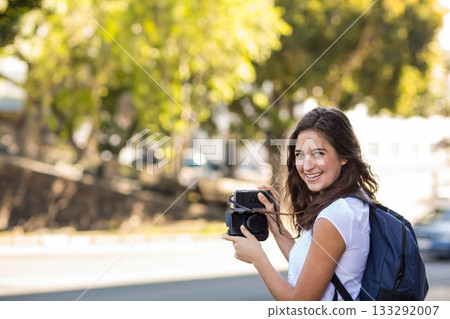 Woman standing beside urban street holding retro camera with blue backpack looking back, copy space Woman standing beside urban street holding retro camera with blue backpack looking back, copy space 133292007