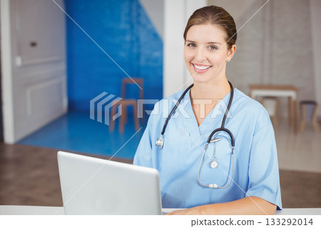 Female nurse wearing scrubs and stethoscope, sitting at desk using laptop in reception, copy space 133292014