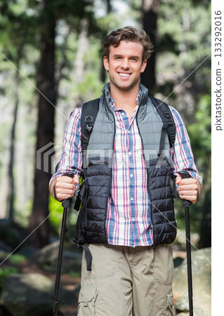 Male hiker wearing quilted vest smiling while trekking pine forest trail with poles and backpack 133292016