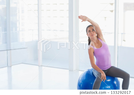 Woman sitting on blue stability ball stretching in fitness studio with city view, copy space Woman sitting on blue stability ball stretching in fitness studio with city view, copy space 133292017