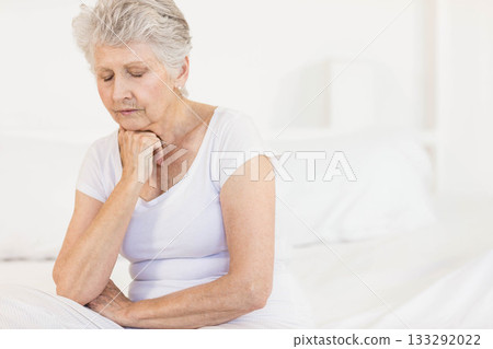 Senior woman sitting on bed edge wearing white pajamas in bright bedroom with headboard, copy space 133292022
