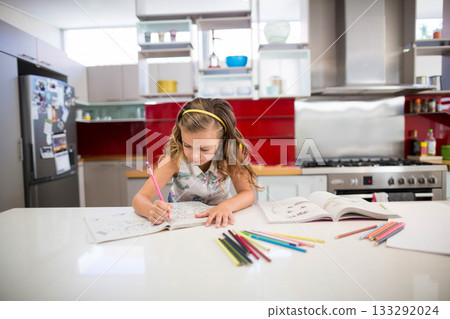 Girl child sitting at white countertop island in kitchen coloring workbook with colored pencils 133292024