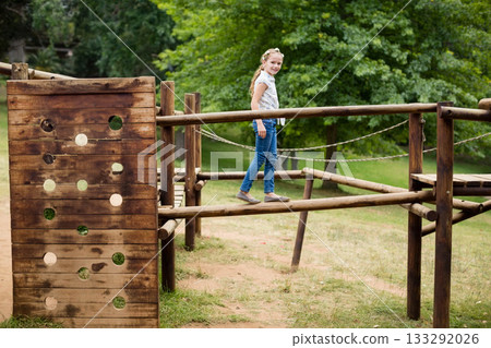 Female child balancing along wooden beam on park playground obstacle course with rope railings 133292026