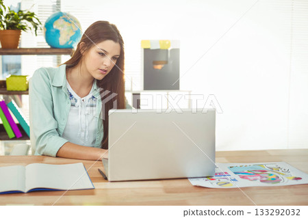 Woman sitting at desk working on laptop with notebook, color printouts in home office, copy space 133292032
