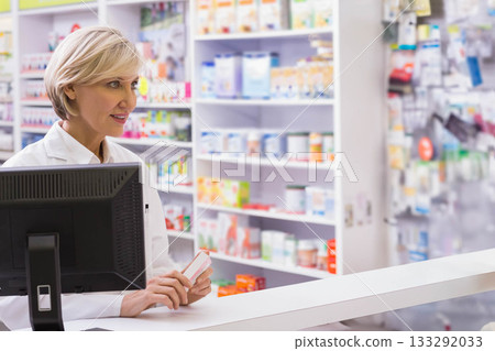 Senior woman pharmacist holding medicine box and checking computer at pharmacy counter, copy space 133292033
