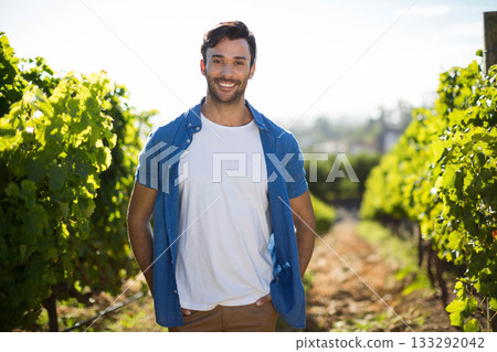 Smiling man standing among vineyard rows and trellis posts on dirt pathway under bright sunlight Smiling man standing among vineyard rows and trellis posts on dirt pathway under bright sunlight 133292042