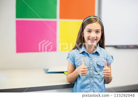 Girl student standing at school desk in bright classroom, holding blue notebook, giving thumbs up 133292048