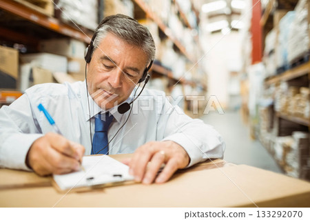 Mature man leaning on carton in warehouse aisle, writing on clipboard with headset Mature man leaning on carton in warehouse aisle, writing on clipboard with headset 133292070