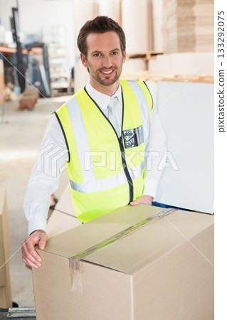 Man wearing neon yellow safety vest holding sealed cardboard box in warehouse with metal shelving 133292075