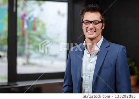 Man standing in office by glass window with ribbons wearing glasses and suit jacket, copy space 133292083