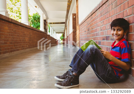 Hispanic boy reading green book while sitting on school corridor floor by brick wall, copy space Hispanic boy reading green book while sitting on school corridor floor by brick wall, copy space 133292089