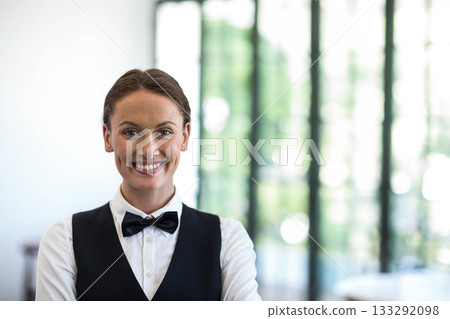 Female server smiling and standing in restaurant lobby wearing white shirt black vest bow tie 133292098