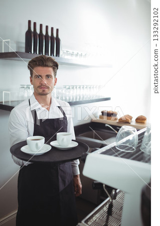 Male barista in white shirt, black apron holding tray with two coffee cups on coffee counter Male barista in white shirt, black apron holding tray with two coffee cups on coffee counter 133292102