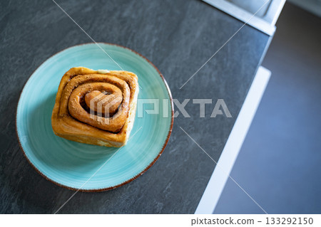 Classic cinnamon roll on an artisan ceramic plate in a home kitchen, top view. Warm tones and natural light create an inviting feel. Concept of breakfast, pastry, comfort, homemade. 133292150