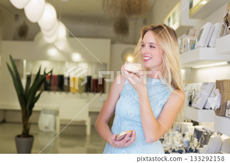 Female shopper with blonde hair sampling scented candle jar with lid by display shelves in boutique Female shopper with blonde hair sampling scented candle jar with lid by display shelves in boutique 133292158