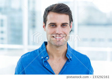Man smiling in blue button-down shirt standing in office with large windows showing urban skyline 133292161