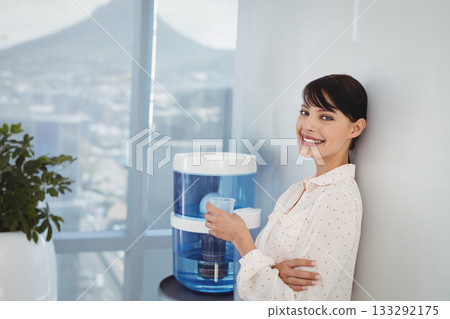 Office woman standing beside blue water cooler holding cup near potted plant at window, copy space 133292175