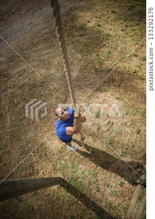 Mid-adult man climbing thick rope on wooden frame under bright sunlight wearing athletic apparel 133292176
