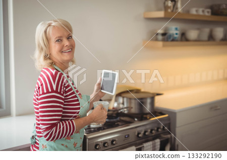 Senior woman in floral apron standing in kitchen holding tablet, mug near stove pot, copy space 133292190