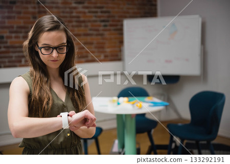 Female in olive dress checking smartwatch on wrist in office with whiteboard and chairs, copy space Female in olive dress checking smartwatch on wrist in office with whiteboard and chairs, copy space 133292191