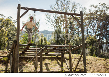 Fit male balancing on wooden platform on outdoor obstacle course with beams, chains, posts Fit male balancing on wooden platform on outdoor obstacle course with beams, chains, posts 133292201