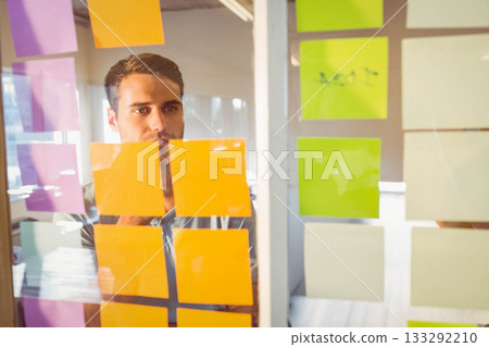 Man in his early thirties reviewing color-coded sticky notes on glass board at open-plan office 133292210