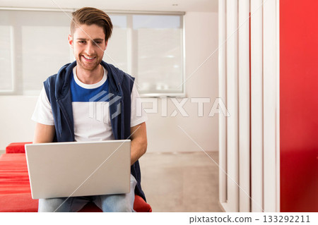 Male wearing navy sleeveless hoodie working on silver laptop on red bench in office lounge smiling 133292211