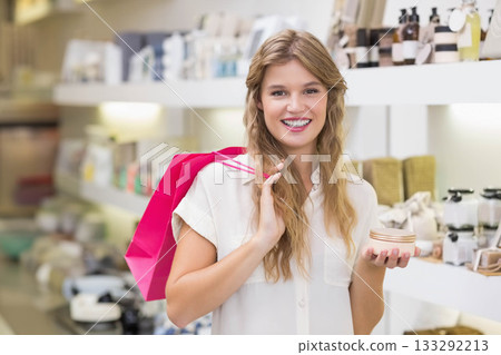Woman smiling while holding cream jar and pink shopping bag in cosmetics store with shelves behind 133292213