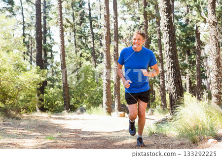 Mature adult man running along sunlit dirt trail in pine woods wearing wrist tracker, copy space Mature adult man running along sunlit dirt trail in pine woods wearing wrist tracker, copy space 133292225