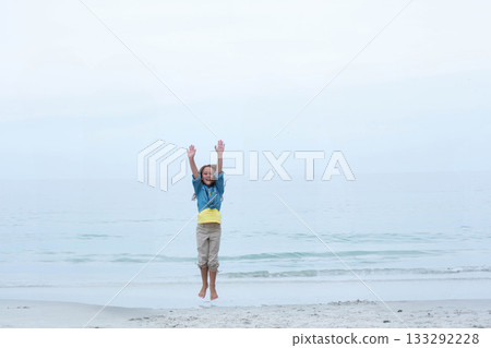 Female child jumping on sandy beach near calm sea under overcast sky wearing denim jacket Female child jumping on sandy beach near calm sea under overcast sky wearing denim jacket 133292228