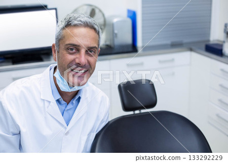 Male dentist leaning on black dental chair in clinic room with monitor, oscillating fan, copy space Male dentist leaning on black dental chair in clinic room with monitor, oscillating fan, copy space 133292229