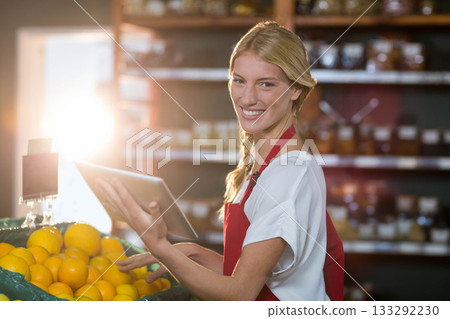 Female grocery clerk wearing red apron using tablet scanning oranges in produce section, copy space 133292230