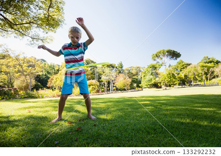 Boy wearing striped polo shirt standing barefoot on grass spinning hula hoop at park, copy space 133292232