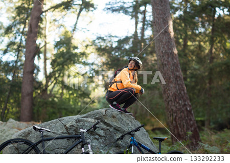 Female cyclist crouching on mossy rock in pine forest adjusting bike helmet beside mountain bikes Female cyclist crouching on mossy rock in pine forest adjusting bike helmet beside mountain bikes 133292233