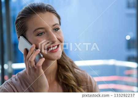 Woman talking on smartphone by glass panel in high-rise interior with neon reflections, copy space 133292237